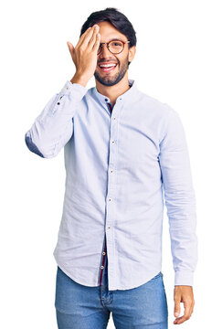 Handsome hispanic man wearing business shirt and glasses covering one eye with hand, confident smile on face and surprise emotion.