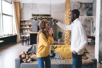 Man in white shirt presents keys to woman in yellow sweater, both beam with joy in spacious, modern...
