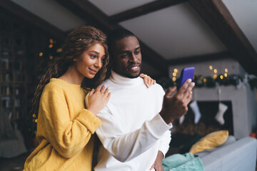 Loving interracial couple engaged with mobile phone; woman in sweater rests her head on man in...