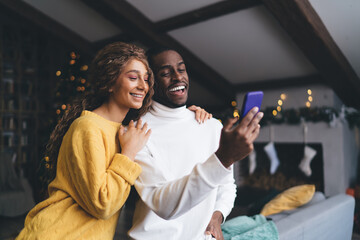 Loving interracial couple engaged with mobile phone; woman in sweater rests her head on man in...