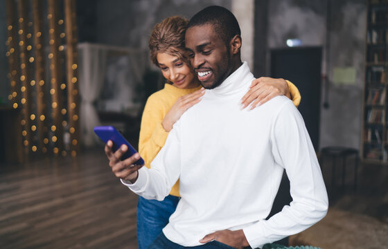 Smiling interracial couple enjoys a moment together; Caucasian woman in a yellow knit hugs from behind an African American man in a white turtleneck, both looking at a smartphone in a warmly lit room - Powered by Adobe
