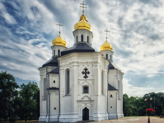 The ancient Orthodox Church of St. Catherine in Chernihiv, Ukraine, graces the scene with its pristine white exterior, golden domes, and crosses, set against the backdrop of a picturesque blue sky.