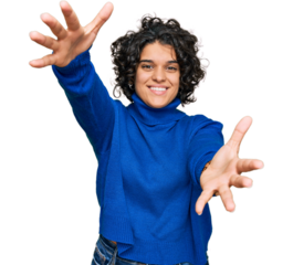 Young hispanic woman with curly hair wearing turtleneck sweater looking at the camera smiling with open arms for hug. cheerful expression embracing happiness.