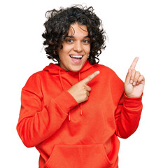 Young hispanic woman with curly hair wearing casual sweatshirt smiling and looking at the camera pointing with two hands and fingers to the side.