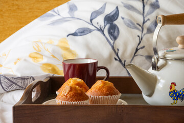 Closeup of tray with colorful teapot, cup and muffins, on a bed with branch patterned pillows, horizontal with copy space