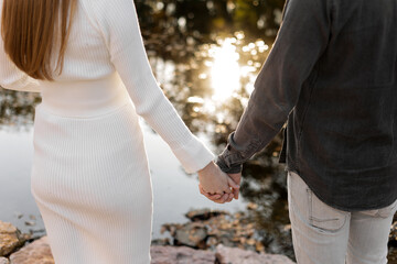Fototapeta premium Rear view of stylish young couple in love holding hands by the pond at autumn park. Woman in knit white dress and hat and man have dating on valentine's day. Happy family love concept