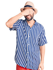Young handsome man with beard wearing summer hat and striped shirt smiling and laughing with hand on face covering eyes for surprise. blind concept.