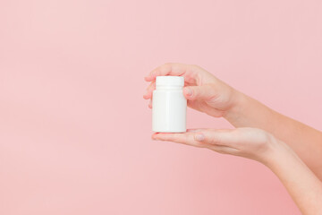 White bottle plastic tube in woman's hands on pink background. Packaging for pills, capsules or supplements. Cosmetics