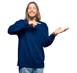 Handsome caucasian man with long hair wearing casual winter sweater amazed and smiling to the camera while presenting with hand and pointing with finger.