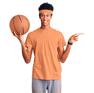Young african american man holding basketball ball smiling happy pointing with hand and finger to the side