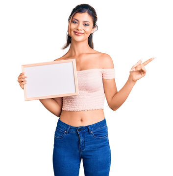 Young beautiful woman holding empty white chalkboard smiling happy pointing with hand and finger to the side