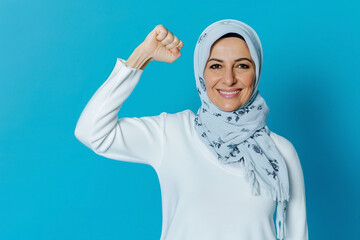 A smiling middle-aged Arab woman standing on a blue background raises her arm, showing her muscles, feeling confident in victory, strong and independent