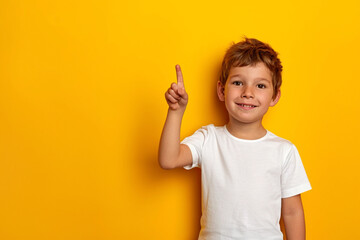 A young boy of Caucasian appearance stands against a yellow wall, pointing and pointing up with his number two finger, smiling a confident and happy smile