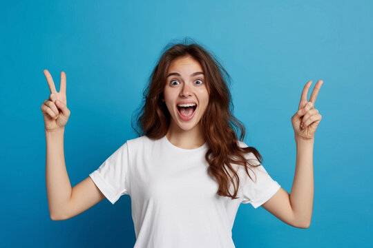A successful, friendly young European brunette in a white T-shirt on a blue background exclaims excitedly, pointing both index fingers up, indicating something