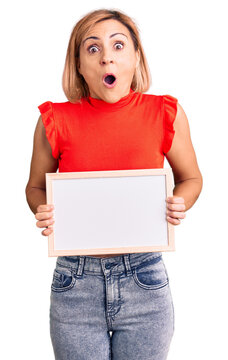 Young blonde woman holding empty white chalkboard scared and amazed with open mouth for surprise, disbelief face
