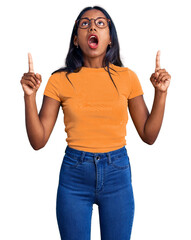 Young indian girl wearing casual clothes and glasses amazed and surprised looking up and pointing...
