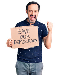 Middle age handsome man holding save our democracy cardboard banner screaming proud, celebrating victory and success very excited with raised arms