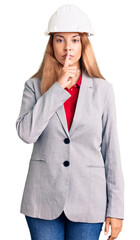 Beautiful young woman wearing architect hardhat asking to be quiet with finger on lips. silence and secret concept.