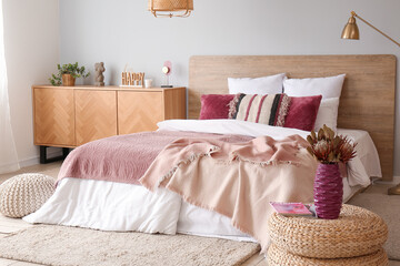 Interior of bedroom with cozy bed, wooden cabinet and dried flowers in vase on wicker pouf