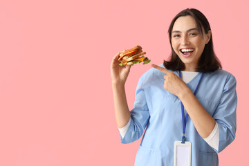 Young female doctor pointing at tasty sandwich on pink background
