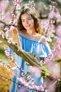 Positive european girl in blue dress standing under blossom peach tree at sunny spring day