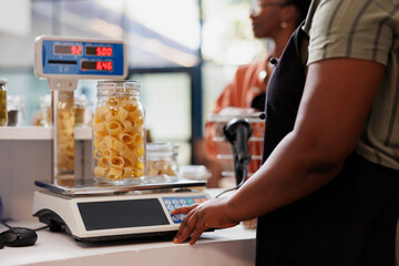 Detailed shot of seller utilizing digital scale to determine weight of container filled with organic produce. Closeup of vendor weighing jar of fresh pasta, with client patiently waiting at counter.