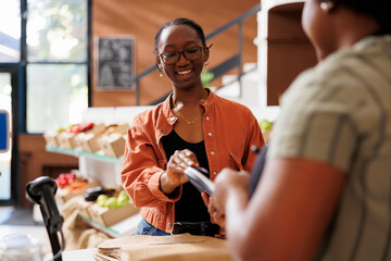 African American woman smiles, interacts with client. Black female customer using credit card on POS for cashless payment. Locally grown organic produce, sustainable farming.