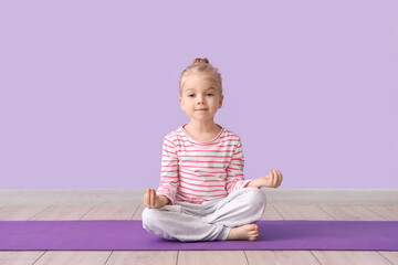 Cute little girl sitting in lotus pose on yoga mat against lilac background