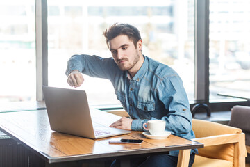 Portrait of angry aggressive young man freelancer in blue jeans shirt working on laptop, having video call, arguing with partner, showing fist to screen. Indoor shot near big window, cafe background.