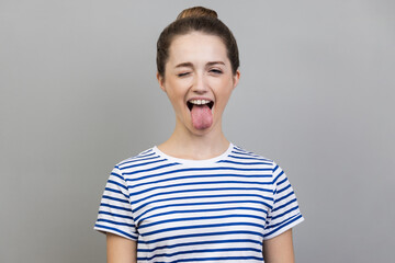 Portrait of disobedient crazy funny woman wearing striped T-shirt showing tongue out and closed her eyes displeased, behaving unruly naughty. Indoor studio shot isolated on gray background. - obrazy, fototapety, plakaty
