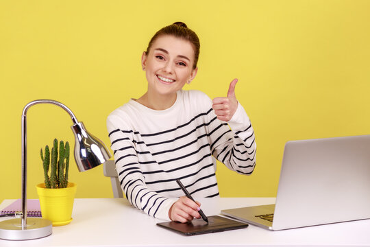 Satisfied smiling woman creative designer sitting at workplace with graphic tablet and laptop, looking at camera and showing thumb up. Indoor studio studio shot isolated on yellow background.