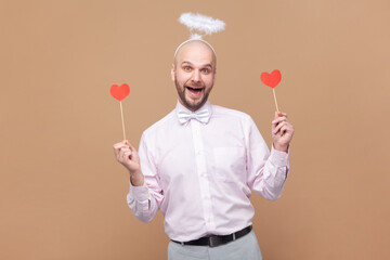 Portrait of happy funny bald bearded man with nimb over head, holding red little hearts, laughing, wearing light pink shirt and bow tie. Indoor studio shot isolated on brown background.
