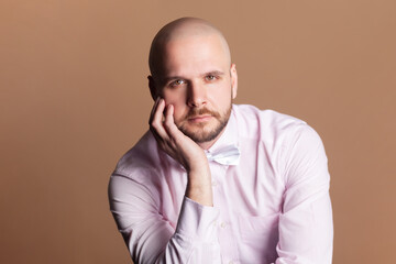 Portrait of calm serious attractive bald bearded man sitting holding his chin, looking at camera, wearing light pink shirt and bow tie. Indoor studio shot isolated on brown background.