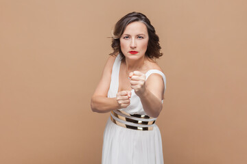 Portrait of angry middle aged woman with wavy hair with clenched fists, fighting with somebody, boxing with anger, wearing white dress. Indoor studio shot isolated on light brown background.