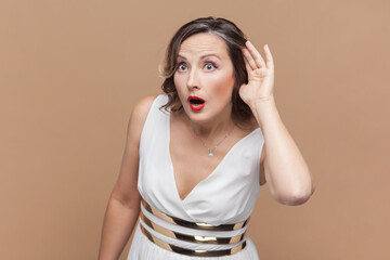 Fototapeta premium Portrait of amazed nosy woman holding hand near ear, listening to interesting talks and private secrets, spying, wearing white dress. Indoor studio shot isolated on light brown background.