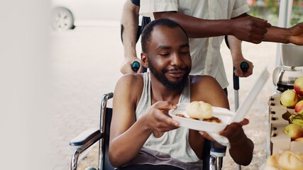 At outdoor food bank, close-up of poor male wheelchair user receiving free food. Volunteers serving...