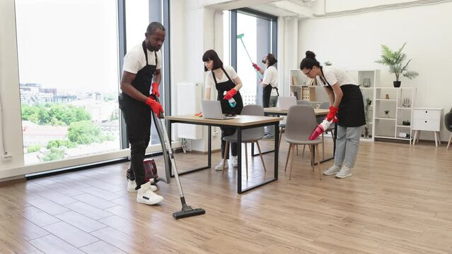 Team of young multicultural cleaners vacuums floor, wipes tables and gadgets, shelves, washes windows in spacious, bright, modern office. Professional cleaning company.