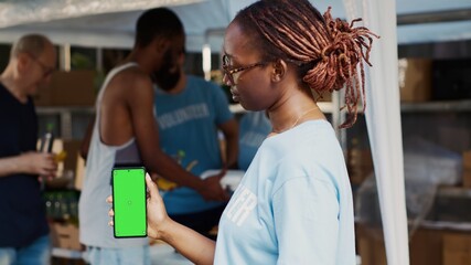 Close-up of african american lady vertically grasping mobile device having blank copyspace chromakey template. During charitable food bank black woman volunteer holds cellphone with green screen. - obrazy, fototapety, plakaty