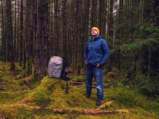 Naklejka premium Male tourist with backpack covered with rain cover in a dense forest. Man in blue jeans and jacket and yellow warm hat. Travel and explore nature concept. Outdoor trip theme.