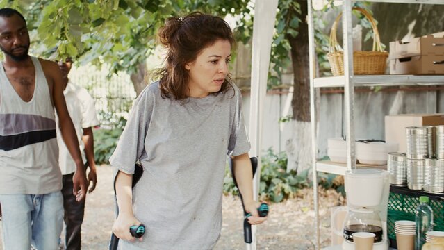 Poor, hungry Caucasian lady on crutches approaches an outdoor food bank to receive free food assisted by black woman. Volunteers serving meals and nourishments to underprivileged and homeless people.