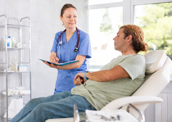 Friendly female doctor holding document folder and engaged in conversation with male patient lying on examination couch in medical office, discussing procedure details or treatment options