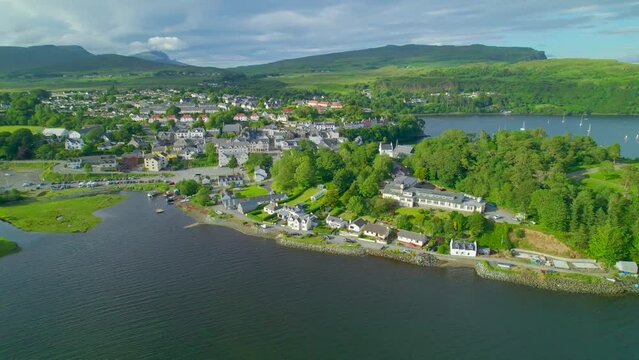 AERIAL: Peaceful bay by the picturesque island town of Portree on a sunny day. Charming port city in sheltered coast of a hilly and green Isle of Skye. Magnificent landscape in northern Scotland.
