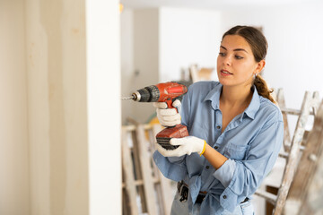 Woman drilling hole in wall with screwdriver in a repairable room - obrazy, fototapety, plakaty