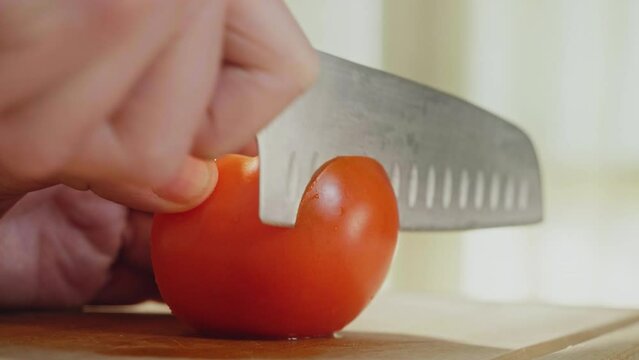 Close up shot of a chef's hands cutting a fresh tomato with knife on wooden chopping board. Cooker preparing vegetarian homemade salad in kitchen, 4k slow motion footage