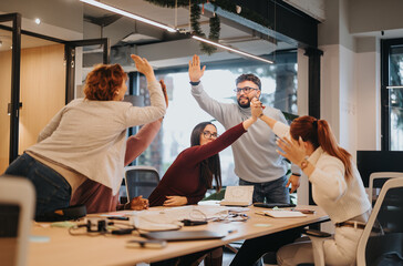 Obraz premium Multicultural colleagues celebrating a successful project in the office. Business people caught up in a moment of victory, smiling and overjoyed with positive outcomes.