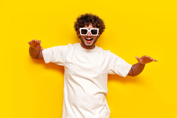 young curly indian guy in sunglasses and white t-shirt dancing and raising his arms on yellow...