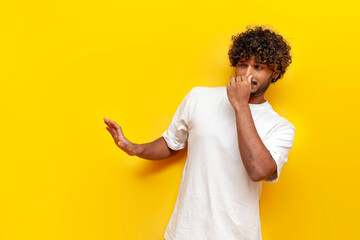 young curly indian man showing disgust and covering his nose with hand on yellow isolated...