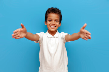 African American boy in white polo wants to hug and pulls his hands forward on blue isolated background, 10 year old child holds empty hands in front of him © Богдан Маліцький