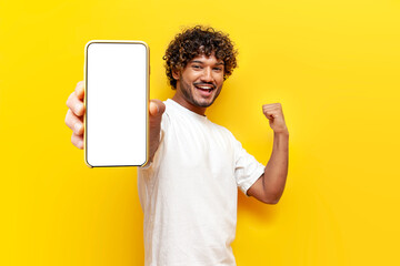 young curly indian man shows blank smartphone screen and rejoices in victory on yellow isolated...