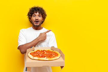 young surprised indian guy holding box of delicious pizza and pointing with his hand to the side on...